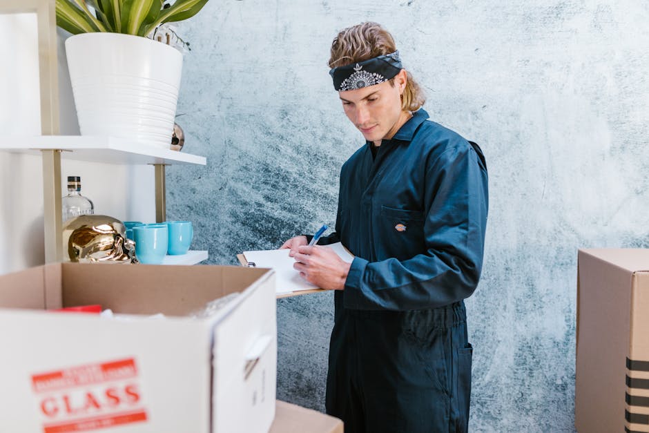 A young male mover from Movers Brent, dressed in a dark blue uniform and wearing a black bandana, is inside a residential property during a home relocation process. He is holding a clipboard and pen, 