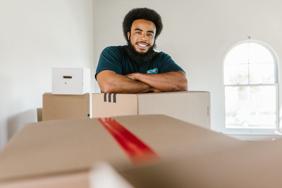 A young male mover from Movers Brent, dressed in a dark blue uniform and wearing a black bandana, is inside a residential property during a home relocation process. He is holding a clipboard and pen, 