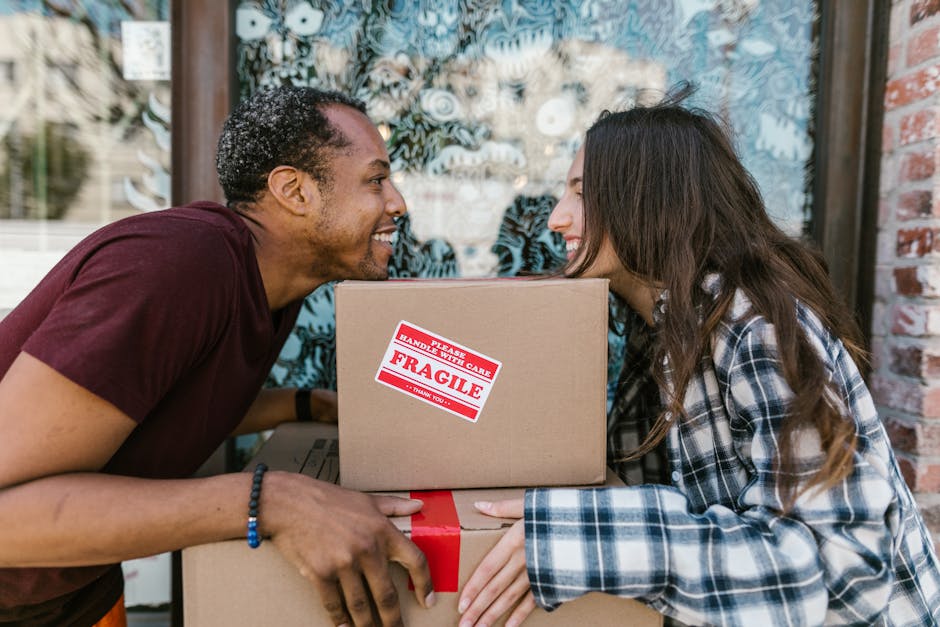 A man and a woman are engaged in a home relocation process outside a building with a glass window decorated with circular patterns. The man, wearing a maroon t-shirt and a beaded bracelet, is leaning forward and smiling at the woman while holding a cardboard box. The woman, dressed in a checkered shirt with long, wavy hair, is also smiling and appears to be touching or supporting the box. The box has a red and white 'FRAGILE' label indicating careful handling. Several similar cardboard boxes are stacked in front of them, with some wrapped in plastic or packing paper. A dolly or trolley used for transporting heavy items is partially visible. The scene takes place on a paved area with the building’s brick wall partially visible to the right, suggesting an outdoor loading or unloading process typical of furniture transport and packing during a house removal, reflecting the services provided by Movers Brent in helping with home relocation and logistics.