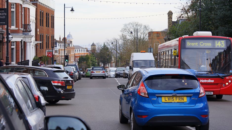 A busy urban street scene showing parked and moving vehicles, including a blue hatchback with the registration plate LV15 FRN, a red Metroline bus displaying route number 143 towards Brent Cross, and several black and white cars lining both sides of the road. The street is lined with brick buildings with shopfronts and residential entrances, lamp posts, and overhead power lines. In the background, trees with budding leaves and overcast sky are visible, indicating a typical day suitable for home relocation and furniture transport. The scene captures the typical traffic and environment encountered during house removals or relocation services, with the presence of a moving van parked along the kerb and other vehicles likely involved in loading or unloading appliances and packed boxes as part of a furniture transport operation by Movers Brent, a company specialising in removals within the Brent area.