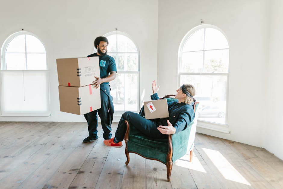 A room during the house removal process showing several cardboard moving boxes of various sizes stacked on top of and beside a dark-colored table with a blue cloth draped over part of it. The boxes ar